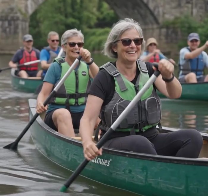 Guests paddling a canoe, laughing, with the old bridge behind them